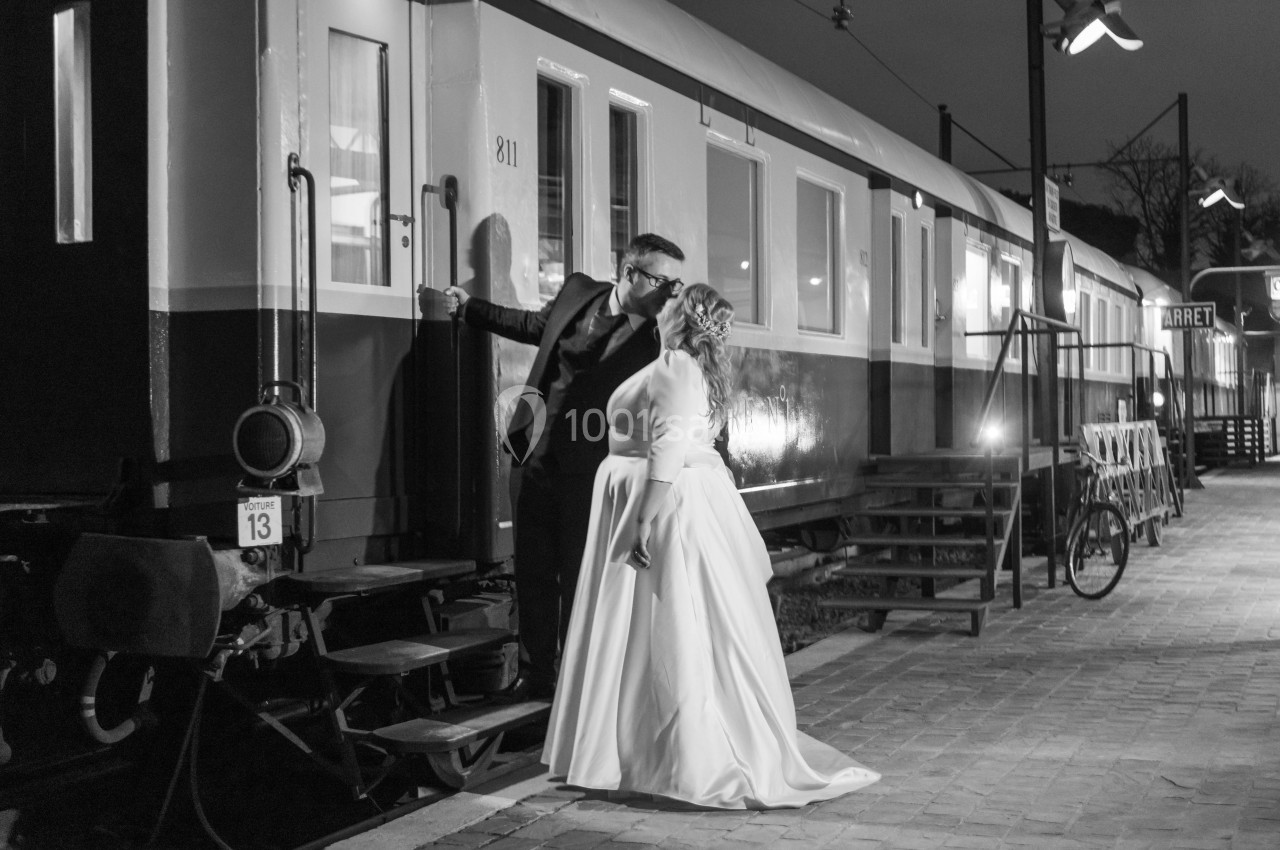 Un couple en tenue de mariage s'embrasse près d'un wagon de train sur un quai éclairé en soirée.