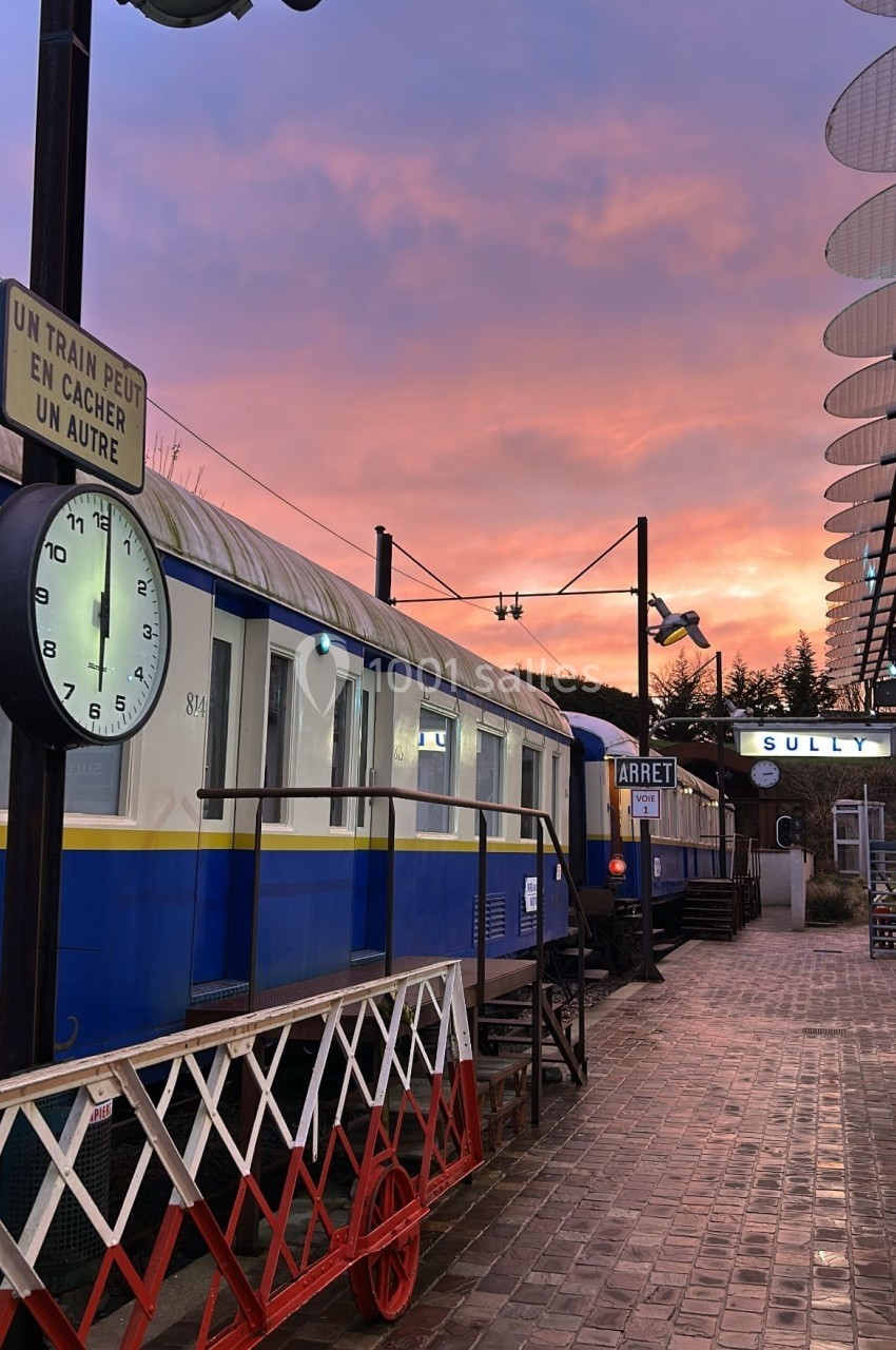 Une gare avec des wagons bleus anciens, une horloge et un ciel coloré au coucher du soleil.