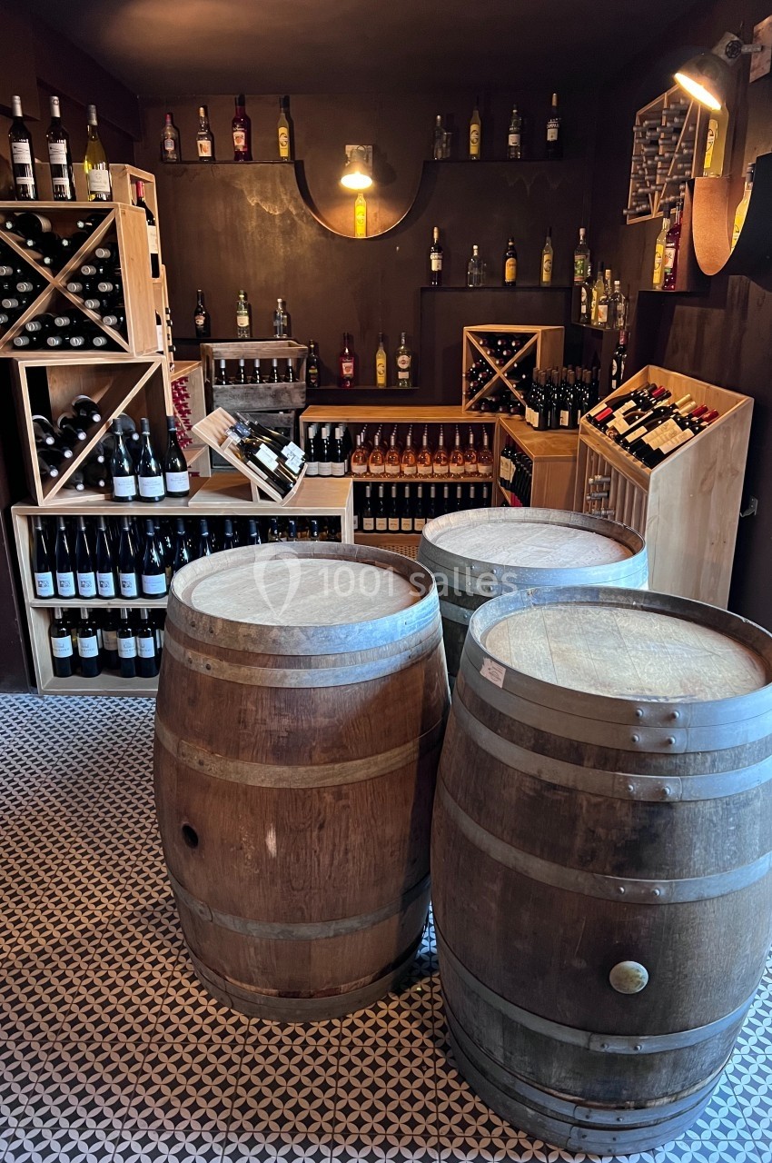Cave à vin avec étagères remplies de bouteilles et trois tonneaux en bois au centre de la pièce.