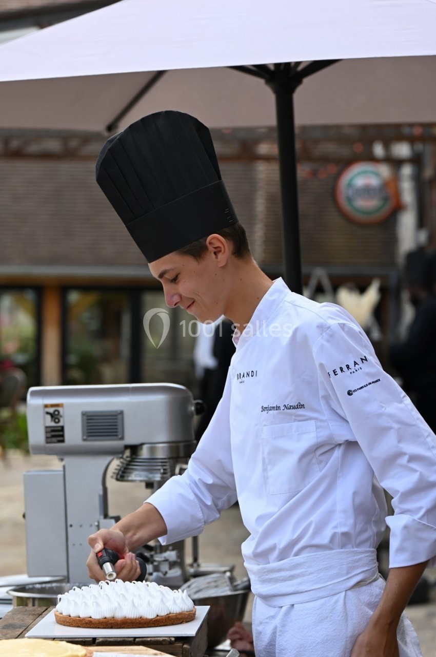 Un chef pâtissier en toque décore une tarte meringuée à l'extérieur, près d'un batteur sur socle.
