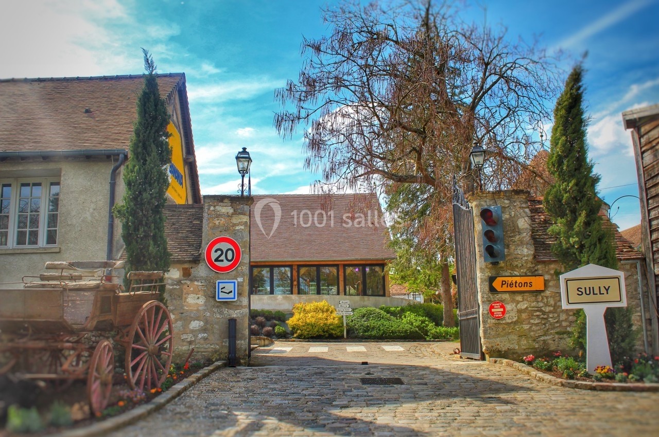 Entrée d'un village avec panneau de limitation à 20 km/h, passage piéton et bâtiments en pierre entourés de végétation.