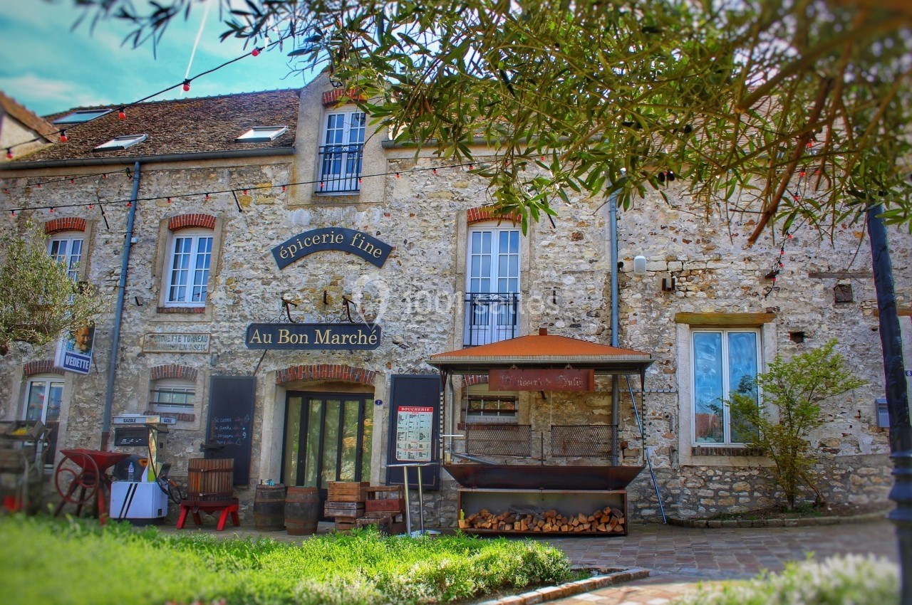 Façade en pierre d'une épicerie fine avec enseigne ’Au Bon Marché’, entourée de végétation et d'éléments rustiques.