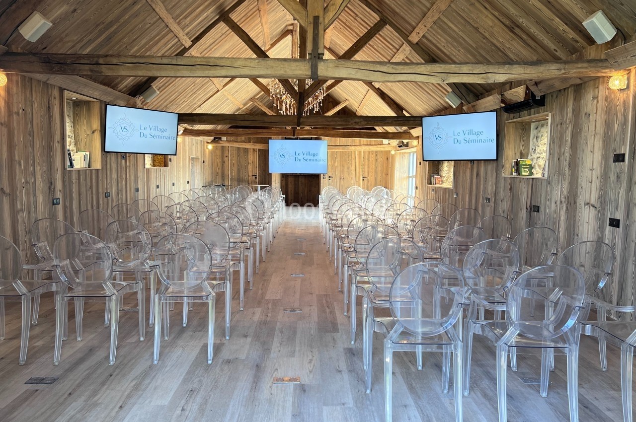 Salle de séminaire en bois avec des chaises transparentes alignées et des écrans affichant ’Le Village du Séminaire’.