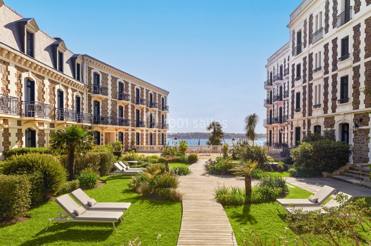 Jardin aménagé avec chaises longues, palmiers et allée en bois, encadré par deux bâtiments en bord de mer.
