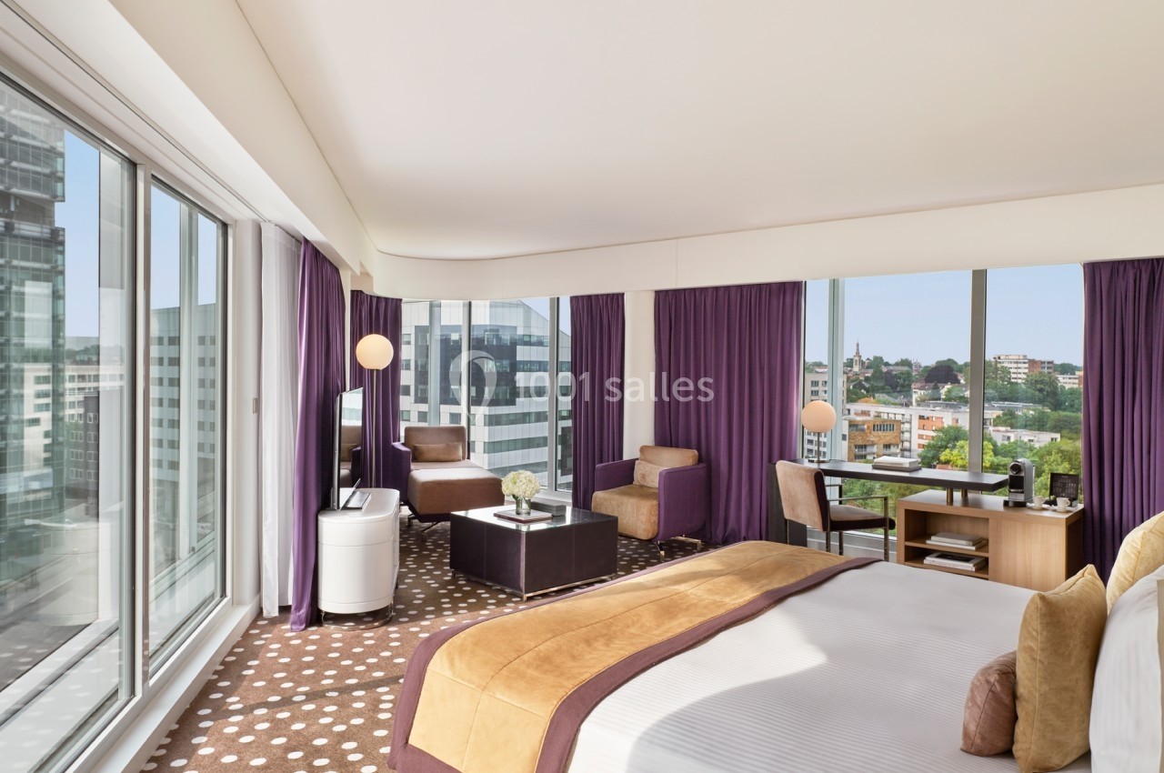 Chambre d'hôtel lumineuse avec lit double, coin salon, bureau et grandes baies vitrées offrant une vue urbaine.