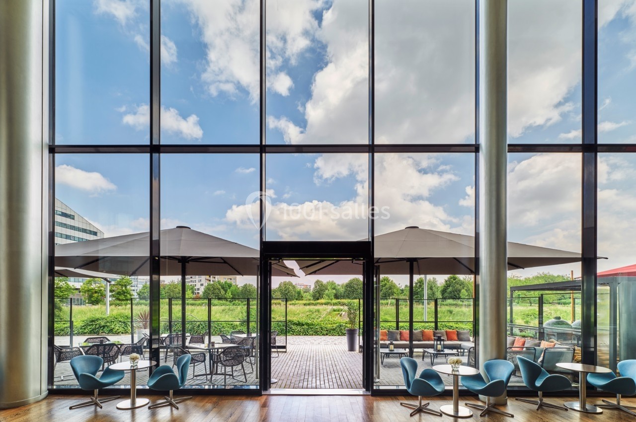 Vue depuis l'intérieur d'un bâtiment avec de grandes baies vitrées donnant sur une terrasse aménagée et un jardin.