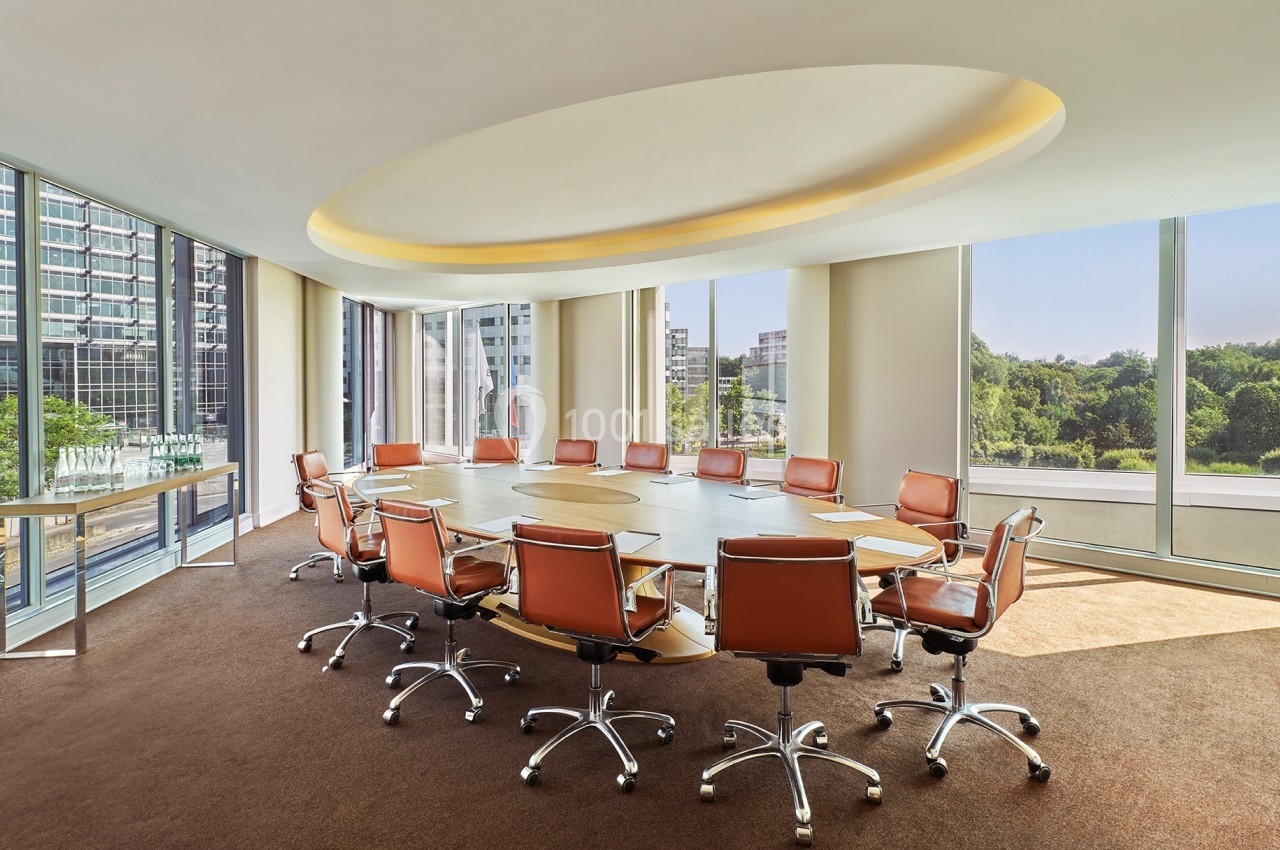 Salle de réunion lumineuse avec une grande table ronde, des chaises en cuir marron et de larges baies vitrées donnant sur l…
