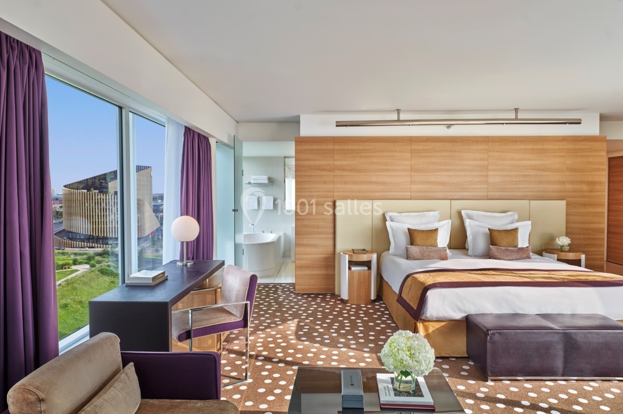 Chambre d'hôtel moderne avec grand lit, bureau, fauteuil, vue sur l'extérieur et salle de bain attenante.