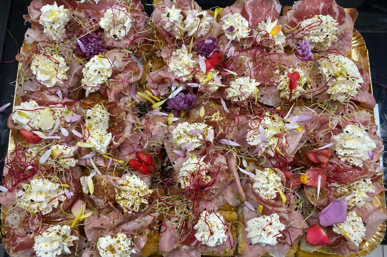 Plateau garni de charcuteries, fromage frais, fleurs comestibles et pousses, disposés sur des tranches de pain.