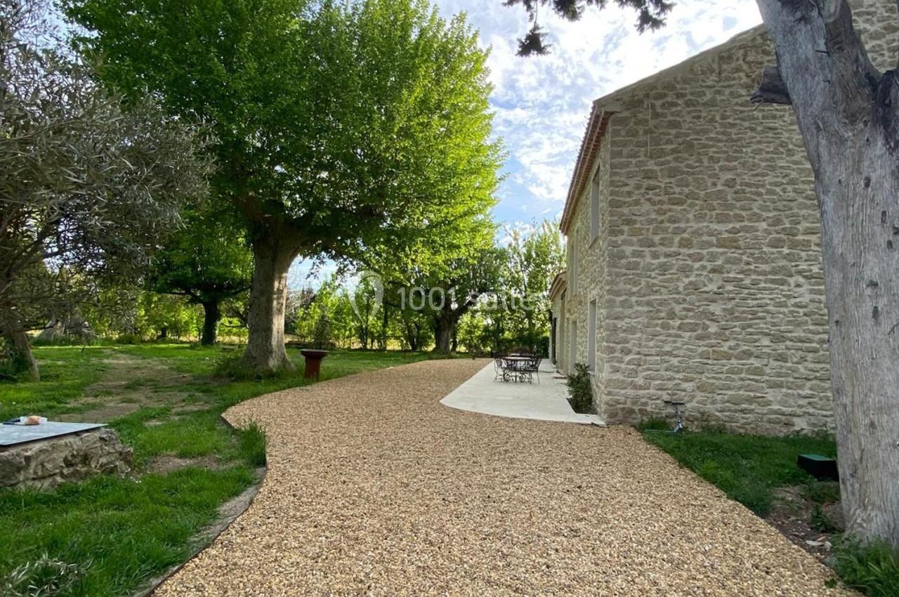 Allée en gravier bordée d'arbres menant à une maison en pierre avec une terrasse aménagée.