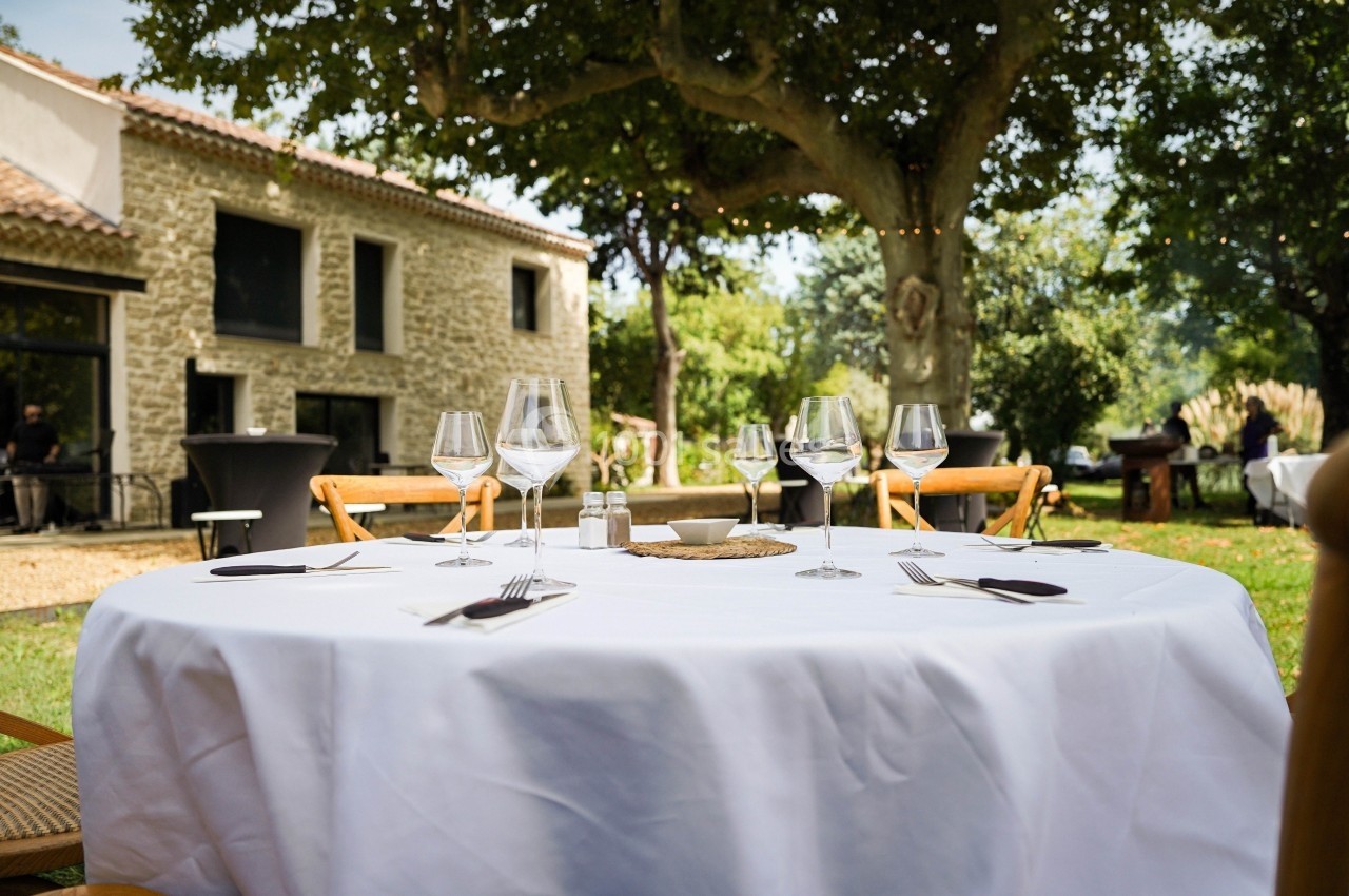 Table dressée en extérieur avec nappage blanc, verres et couverts, devant une maison en pierre et des arbres.