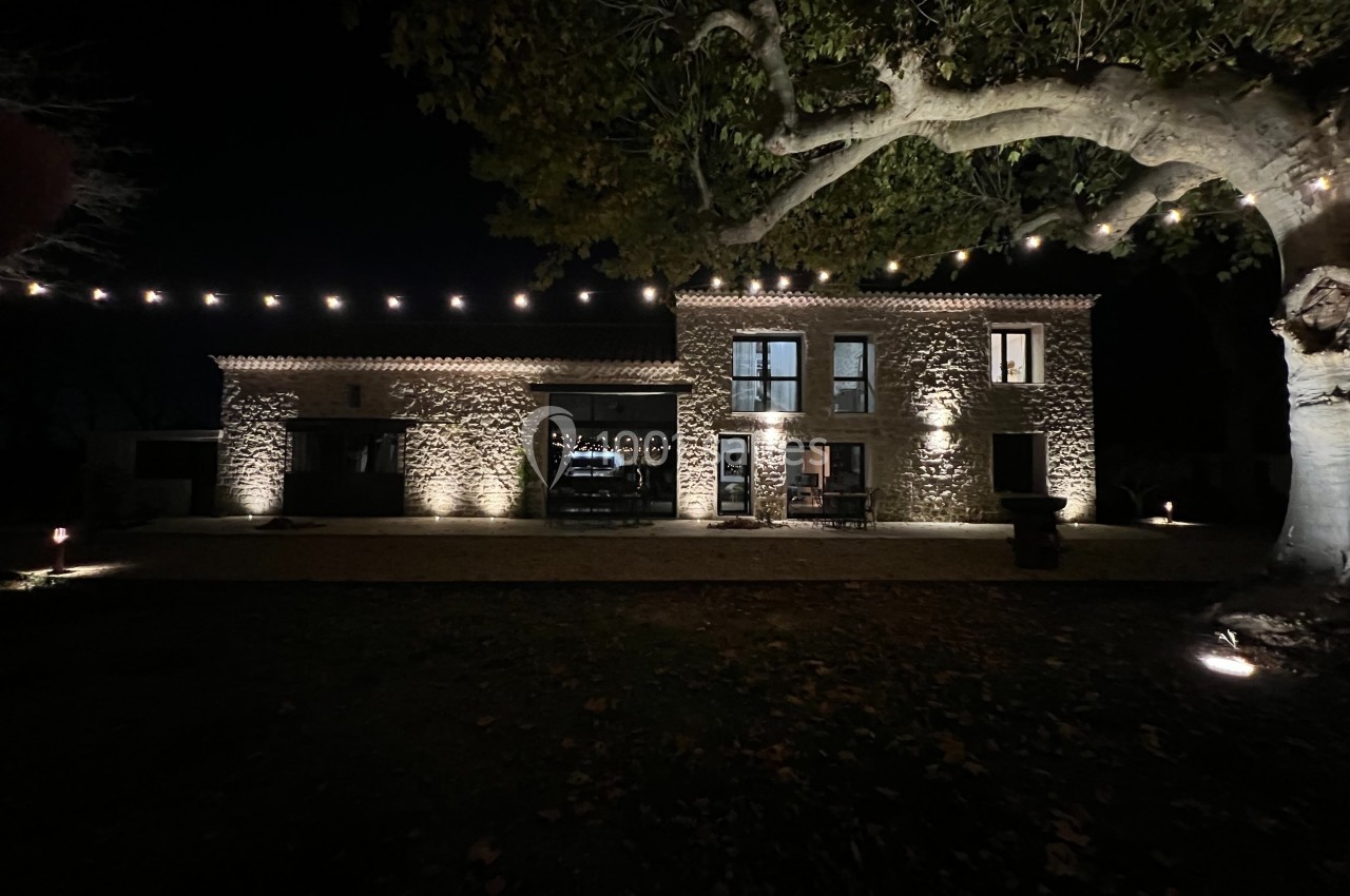 Façade d'une maison en pierre éclairée la nuit, entourée d'arbres et de guirlandes lumineuses suspendues.