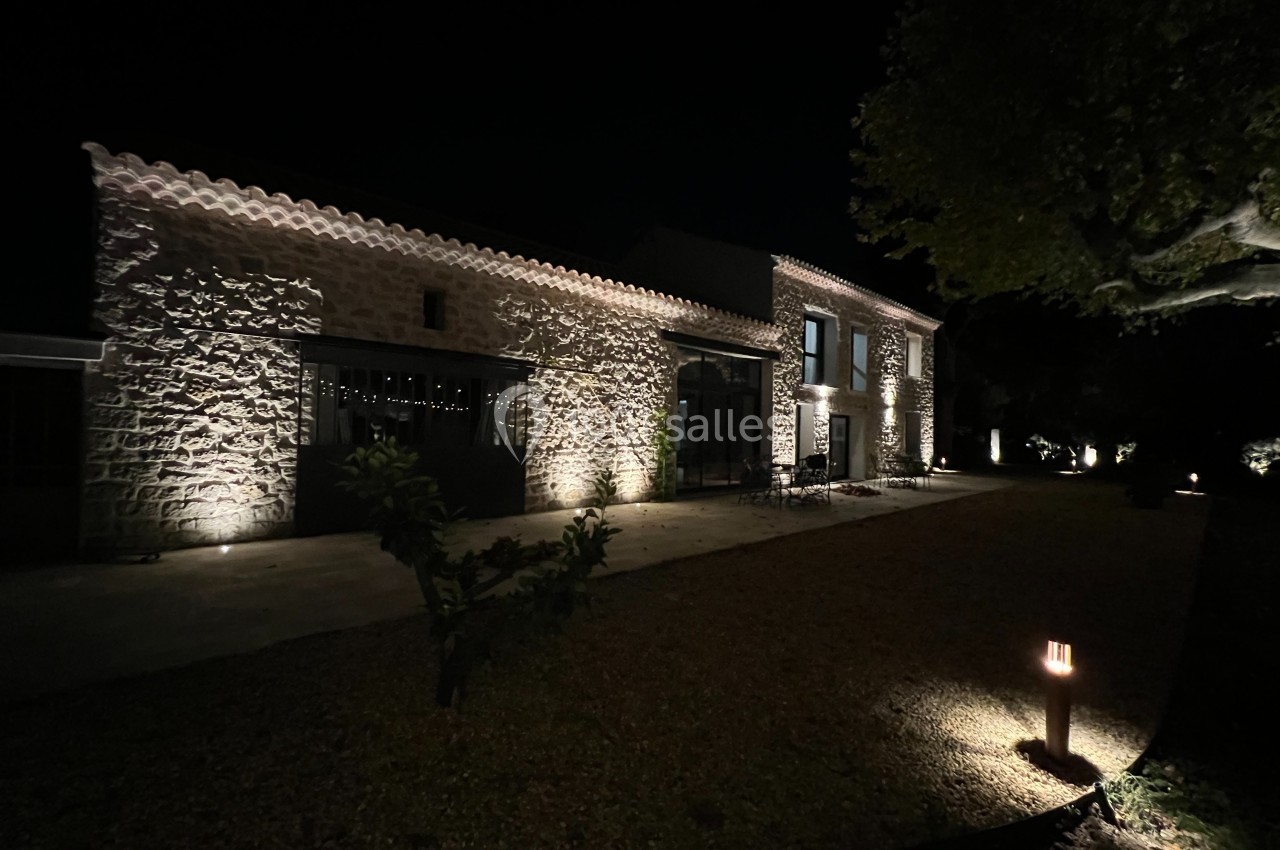 Façade en pierre d'une maison éclairée la nuit, entourée d'un jardin avec un arbre et un chemin gravillonné.