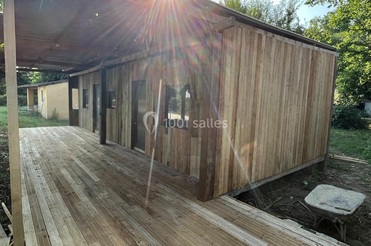Maison en bois avec terrasse en bois, partiellement ombragée, entourée de végétation sous un soleil éclatant.