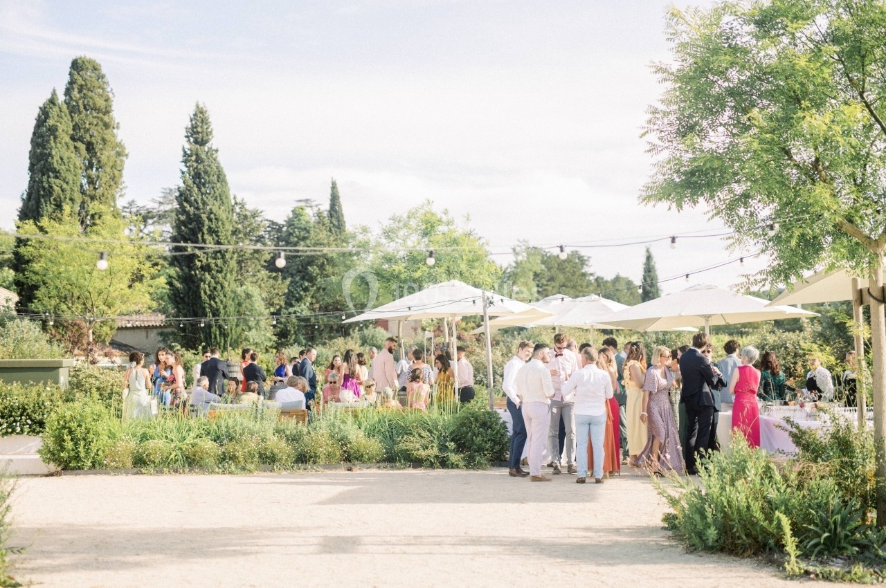 Groupe de personnes rassemblées dans un jardin ensoleillé avec parasols, arbres et guirlandes lumineuses en arrière-plan.