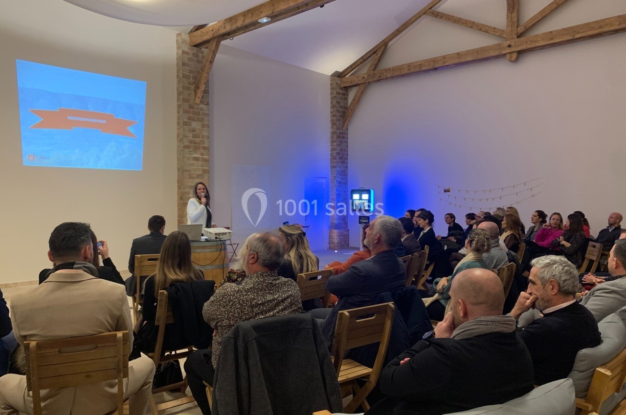 Une conférencière présente devant un public assis dans une salle avec poutres apparentes et écran de projection.