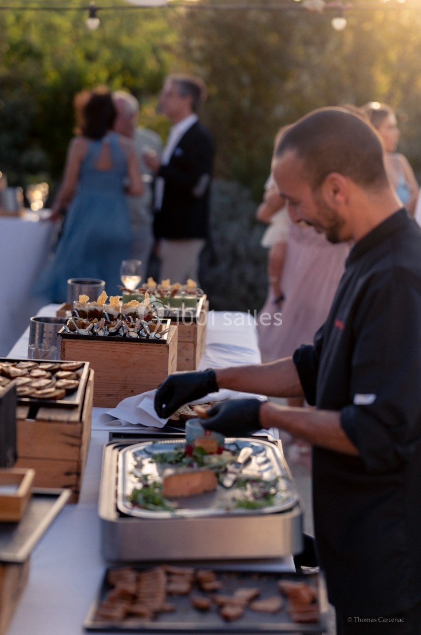 Un chef prépare des plats lors d'un buffet en extérieur, avec des invités en arrière-plan.