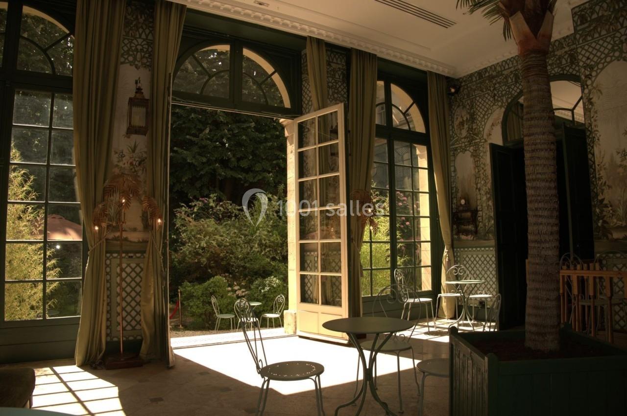 Salle lumineuse avec grandes fenêtres ouvertes sur un jardin verdoyant, mobilier en métal et décorations élégantes.