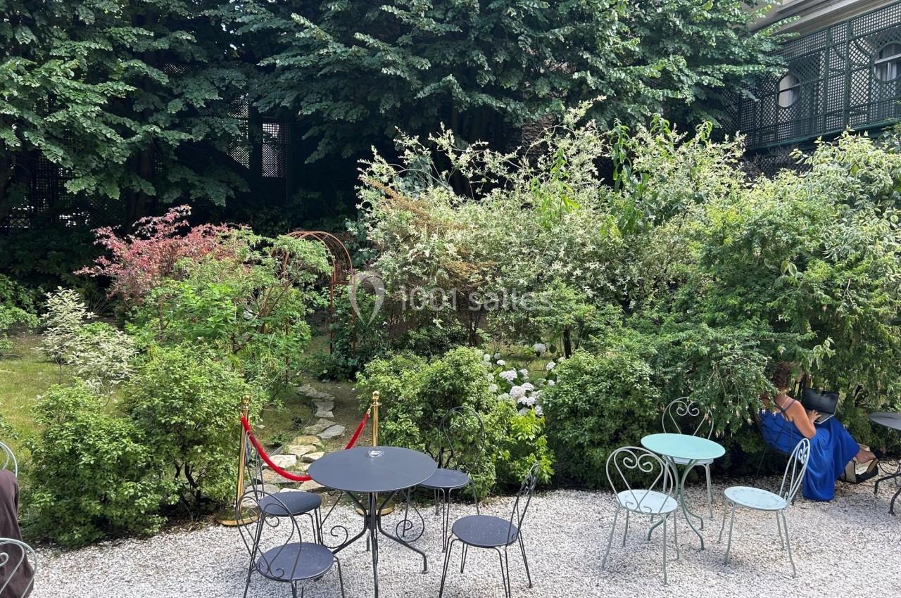 Terrasse avec tables et chaises en métal sur gravier, entourée de végétation dense et d'arbustes fleuris.