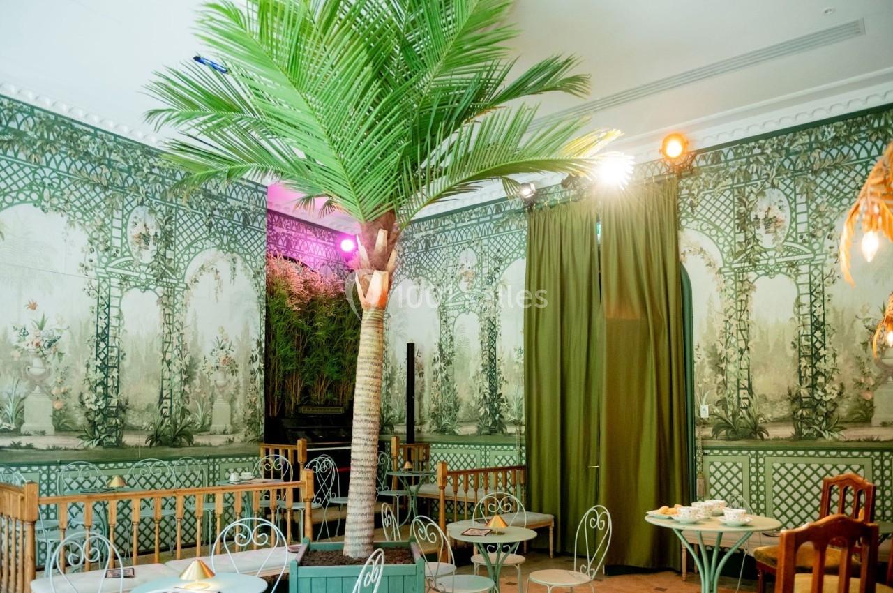 Salle décorée de motifs végétaux, avec un faux palmier, des tables en fer forgé et des rideaux verts.