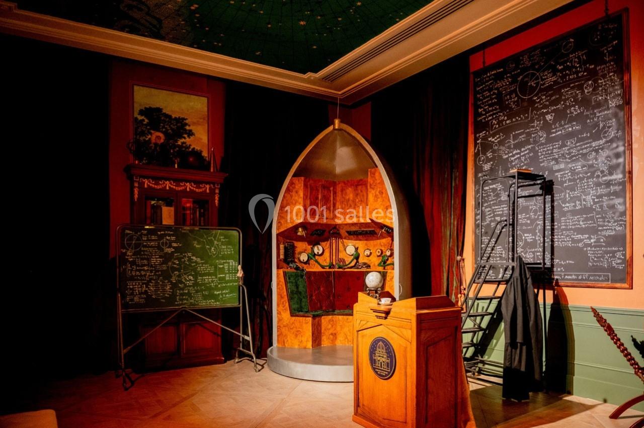 Salle avec un décor scientifique comprenant des tableaux noirs remplis de formules, un pupitre et une capsule futuriste.