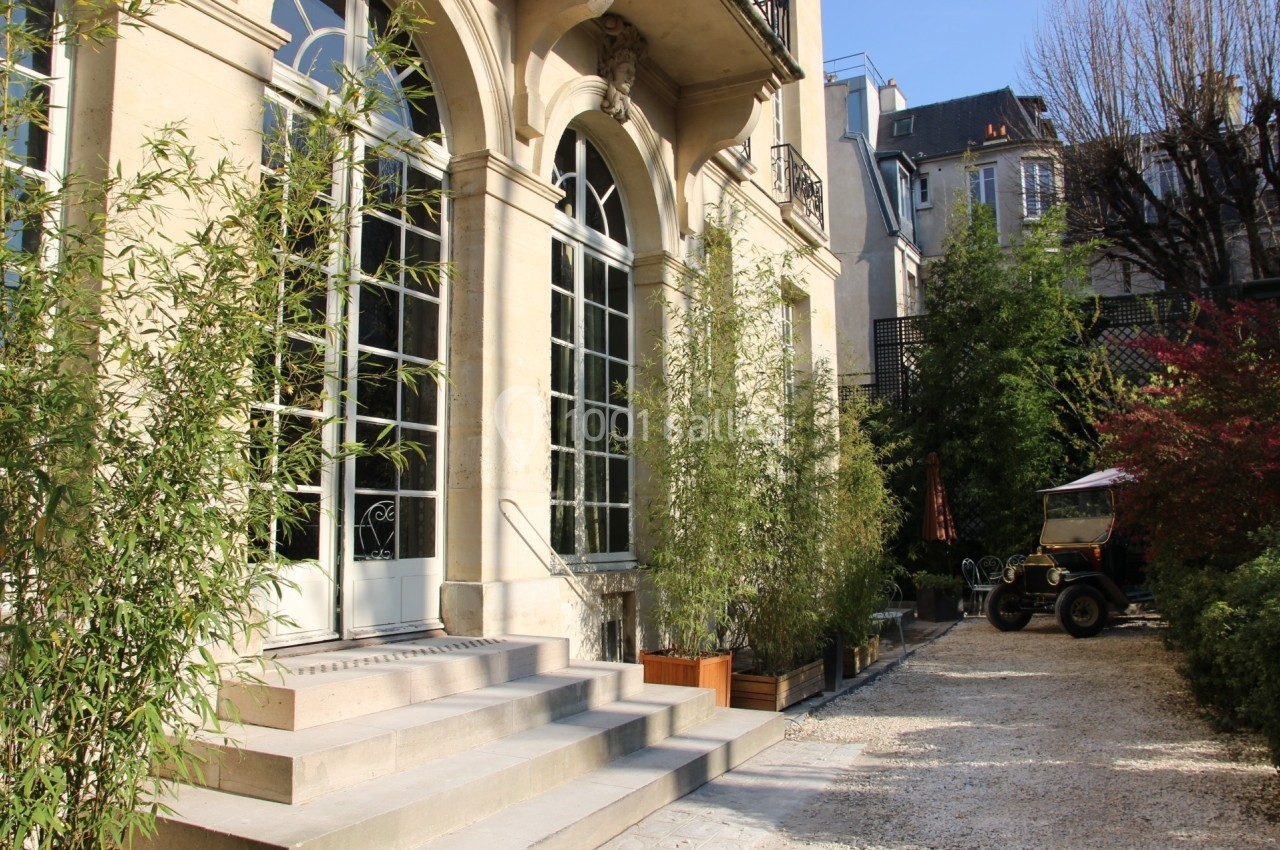 Façade d'un bâtiment ancien avec de grandes fenêtres, entouré de bambous et d'une cour gravillonnée.