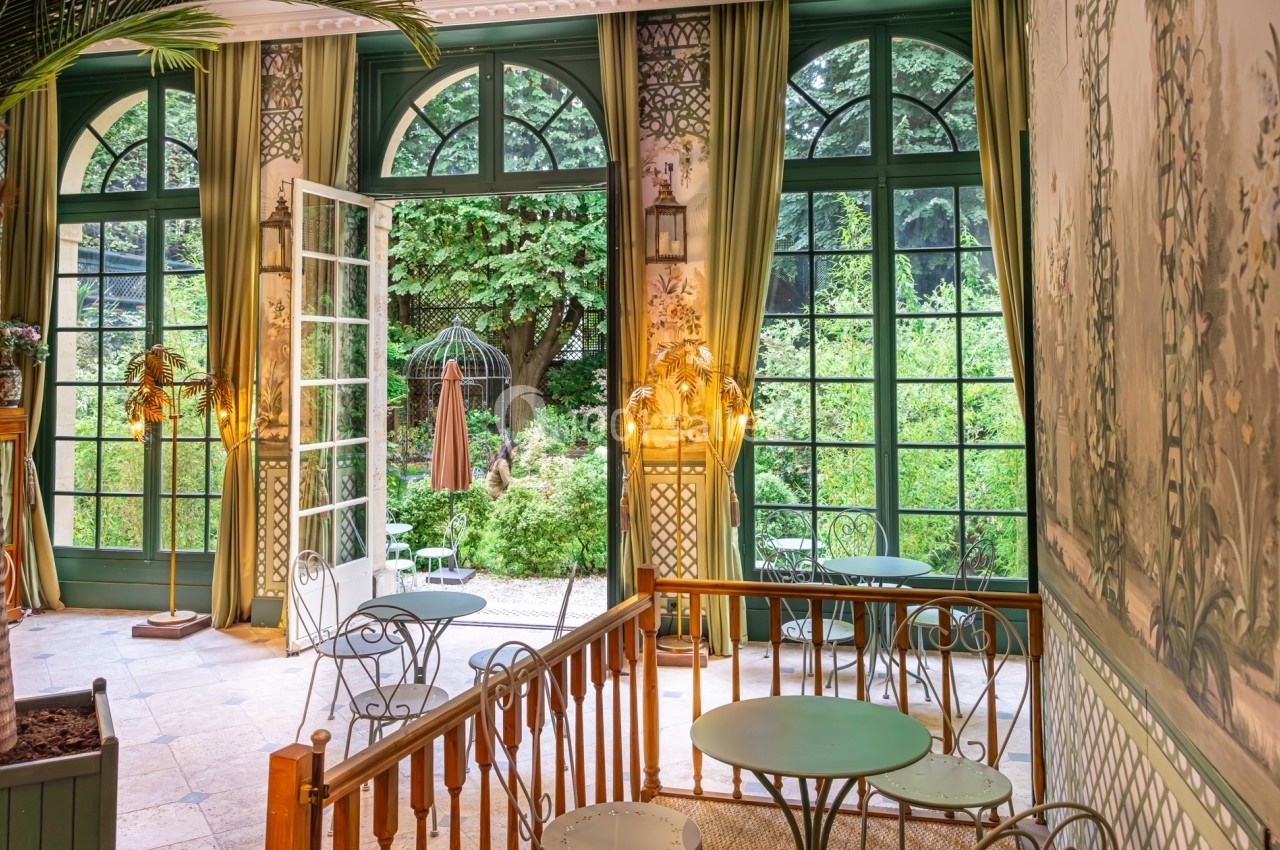 Salle lumineuse avec grandes baies vitrées ouvertes sur un jardin verdoyant, tables et chaises en métal.