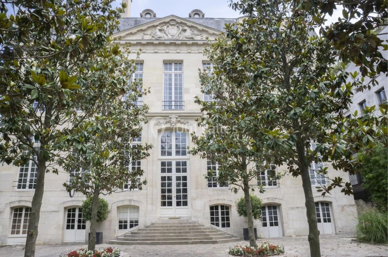 Façade d'un bâtiment en pierre de style classique entouré d'arbres et de parterres de fleurs.
