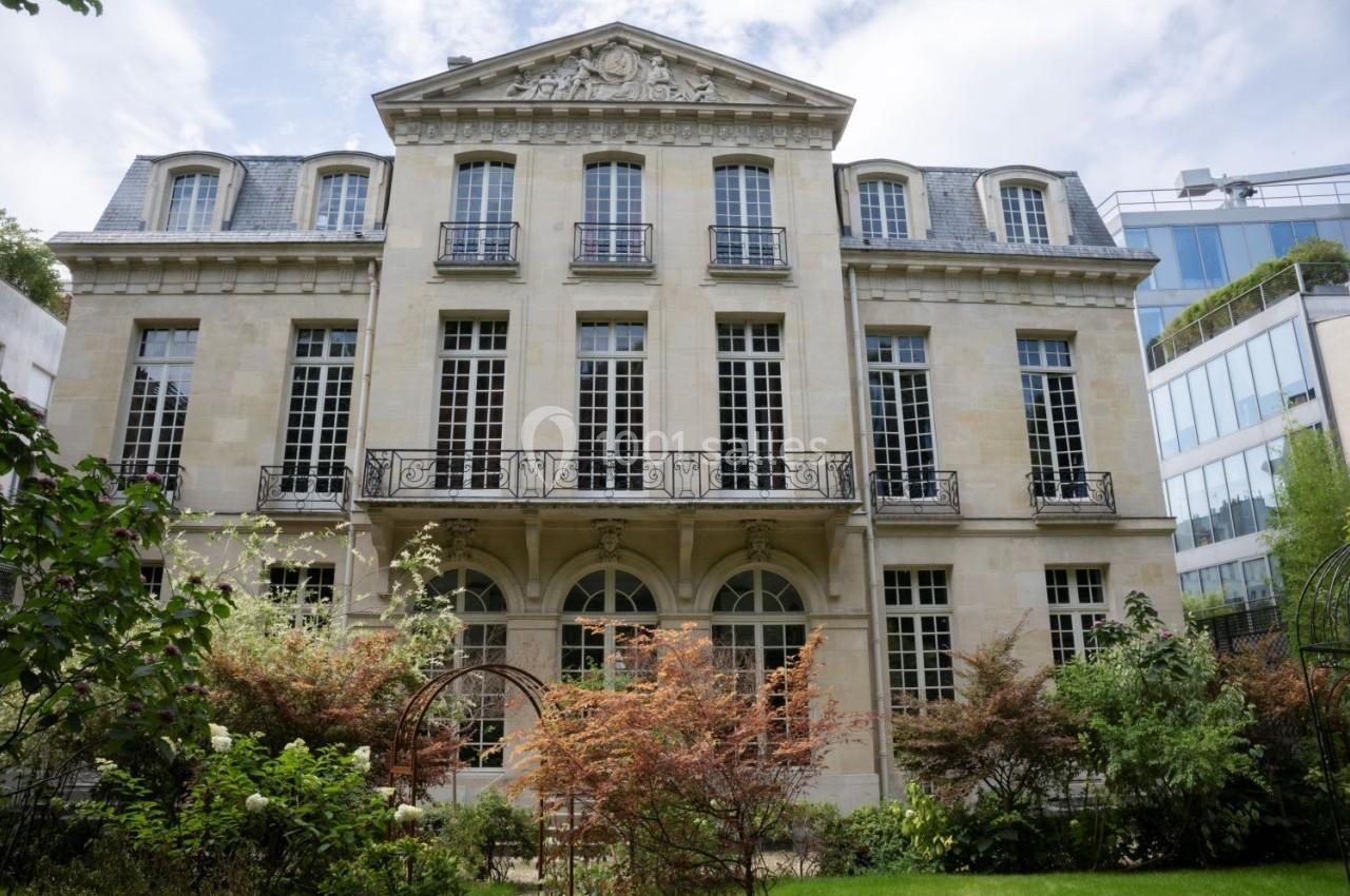 Façade d'un bâtiment ancien en pierre avec grandes fenêtres, entouré de végétation et d'arbustes colorés.
