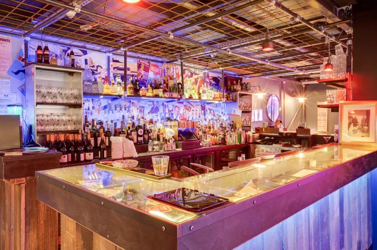 Bar avec comptoir éclairé, étagères remplies de bouteilles, décorations colorées et ambiance lumineuse tamisée.