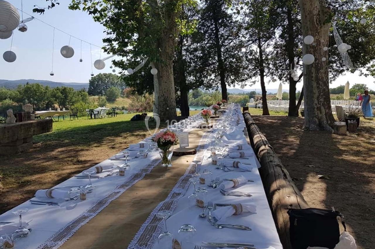Grande table extérieure décorée pour un repas, sous des arbres avec des lanternes suspendues, dans un cadre champêtre.