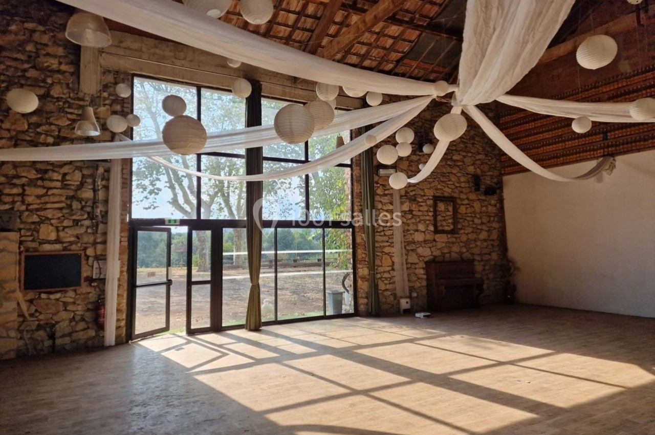 Salle en pierre avec de grandes baies vitrées, décorée de lanternes et de voilages suspendus, éclairée par la lumière…