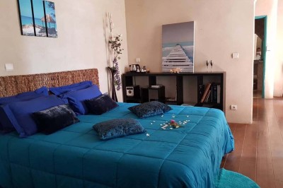 Chambre avec lit double bleu, tête de lit en rotin, décoration sobre et tableau représentant un ponton sur la mer.