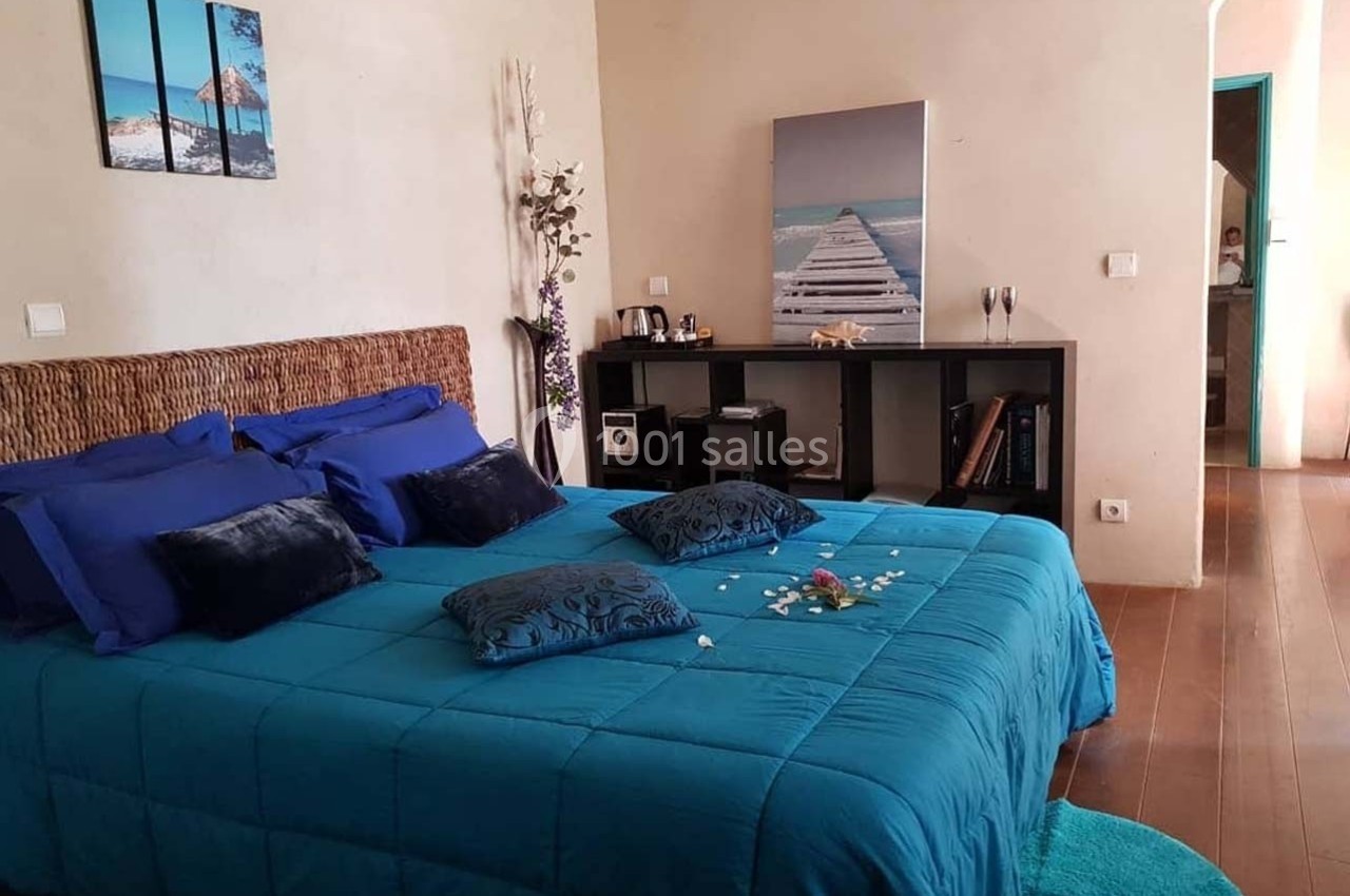 Chambre avec lit double bleu, tête de lit en rotin, décoration sobre et tableau représentant un ponton sur la mer.