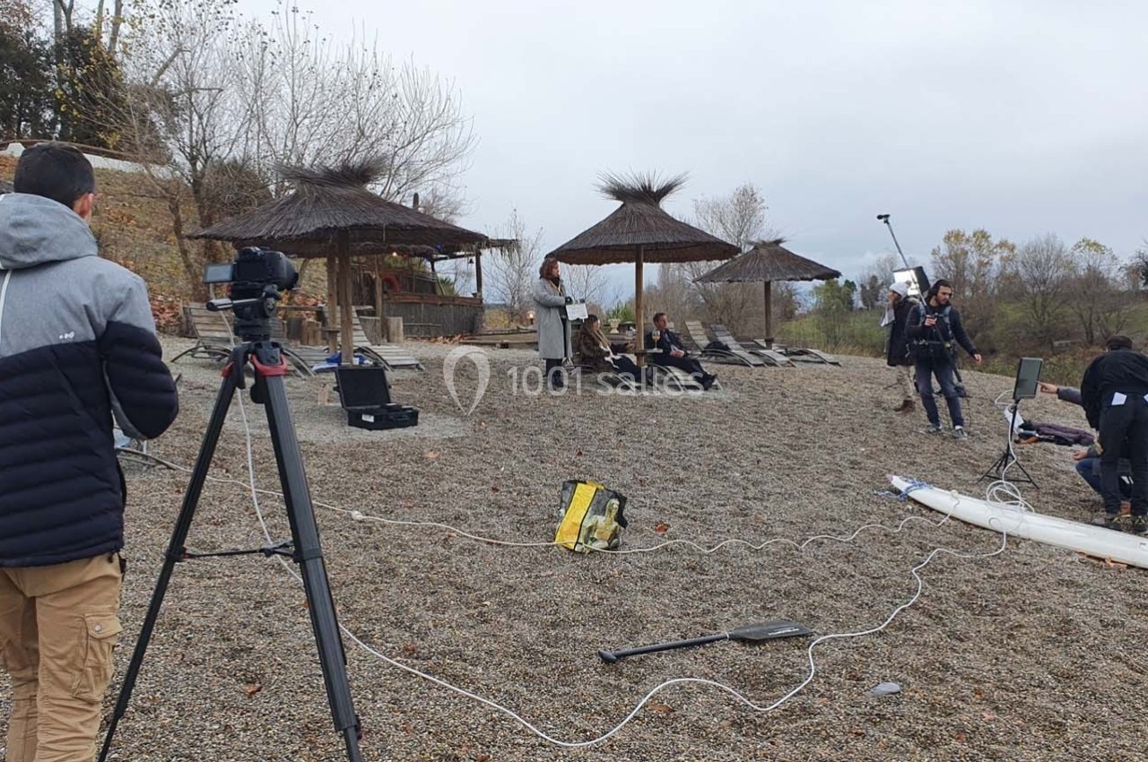 Tournage en extérieur sur une plage avec des parasols en paille, des caméras et une équipe technique en action.