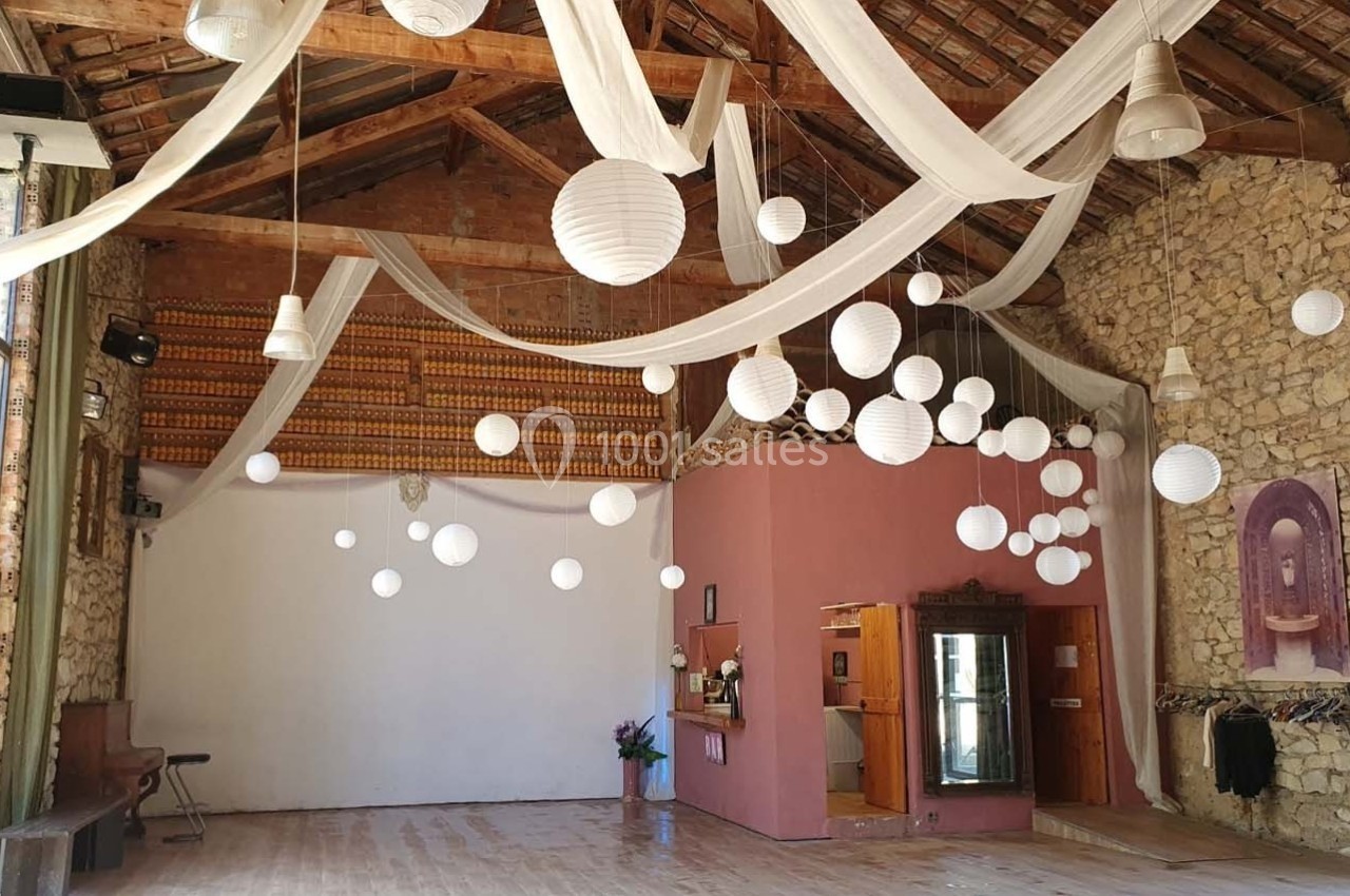 Salle avec murs en pierre, parquet en bois, lanternes suspendues et voilages blancs sous un plafond en poutres apparentes.