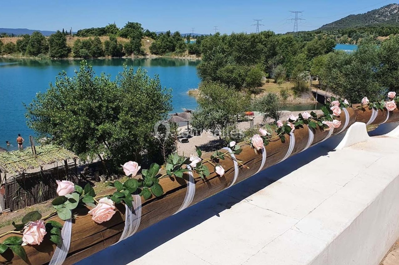 Barrière en bois décorée de roses artificielles roses, surplombant un lac entouré de végétation et de collines.