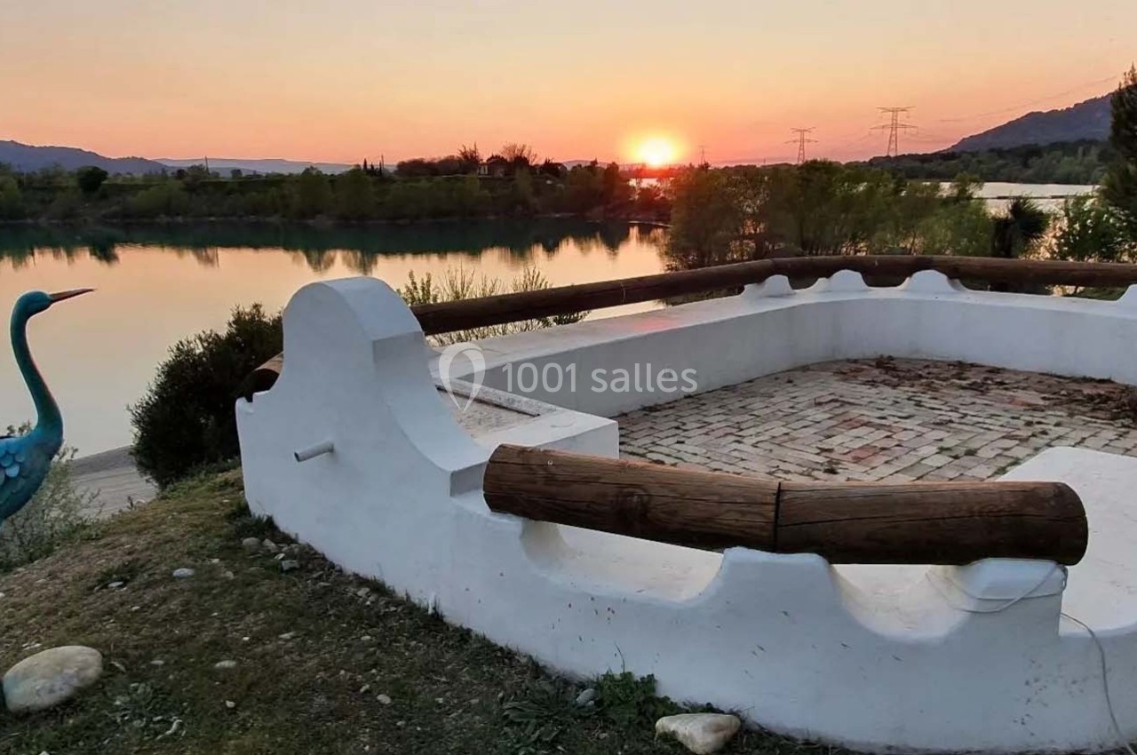 Terrasse en pierre blanche avec vue sur un lac au coucher du soleil, entourée de végétation et d'une statue d'oiseau.