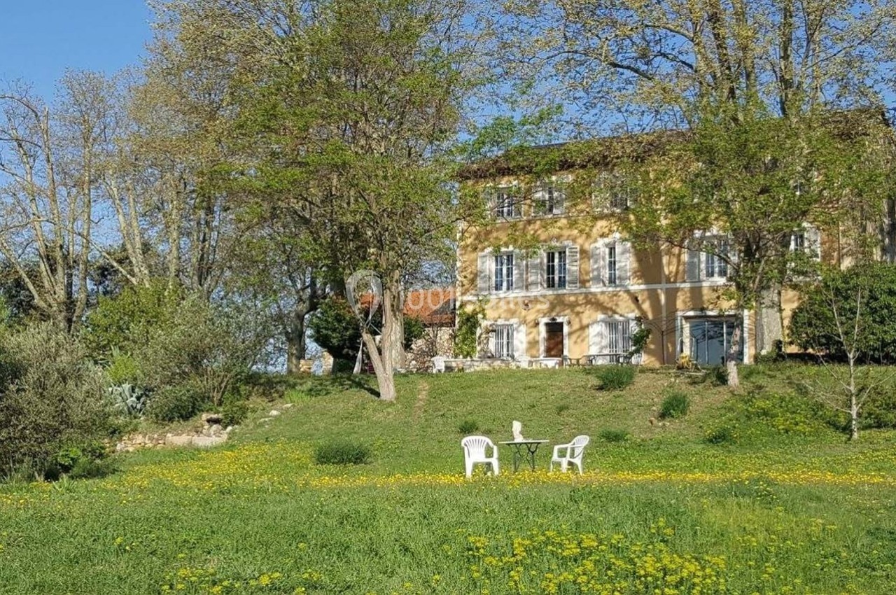 Maison en pierre entourée d'arbres et d'une pelouse fleurie, avec deux chaises blanches et une table au premier plan.
