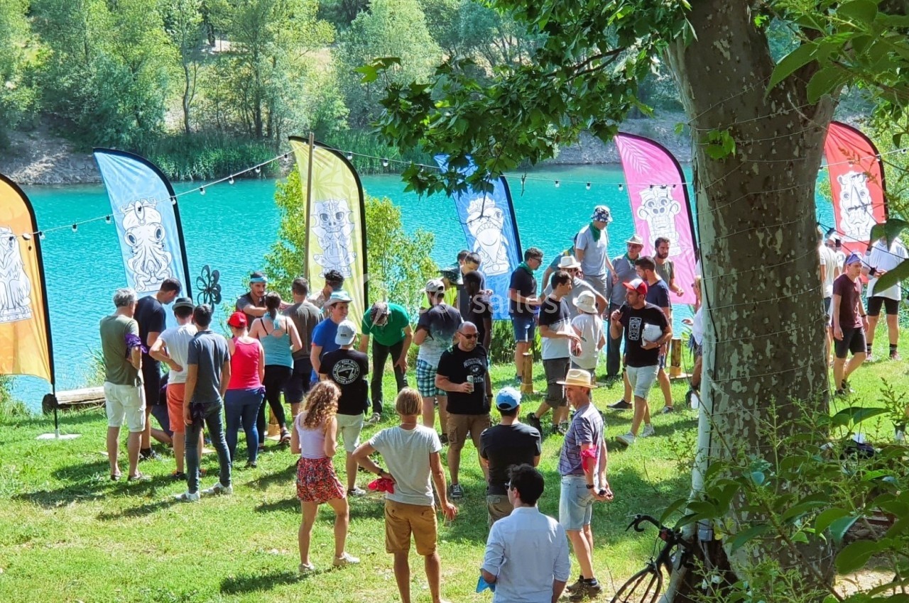 Groupe de personnes rassemblées sur une pelouse près d'un lac, entourées de drapeaux colorés et d'arbres.