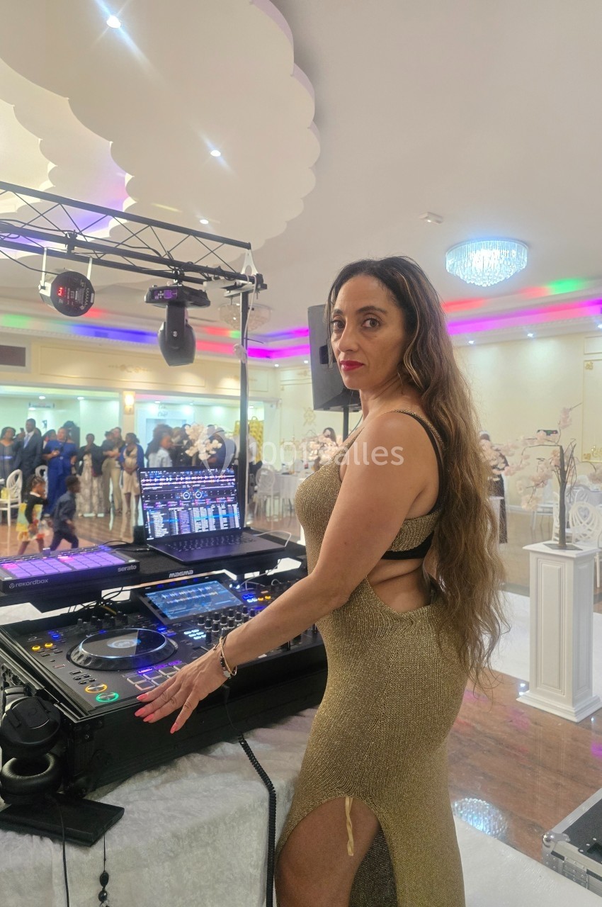 Une femme en robe dorée se tient près d'une table de mixage dans une salle de réception éclairée.