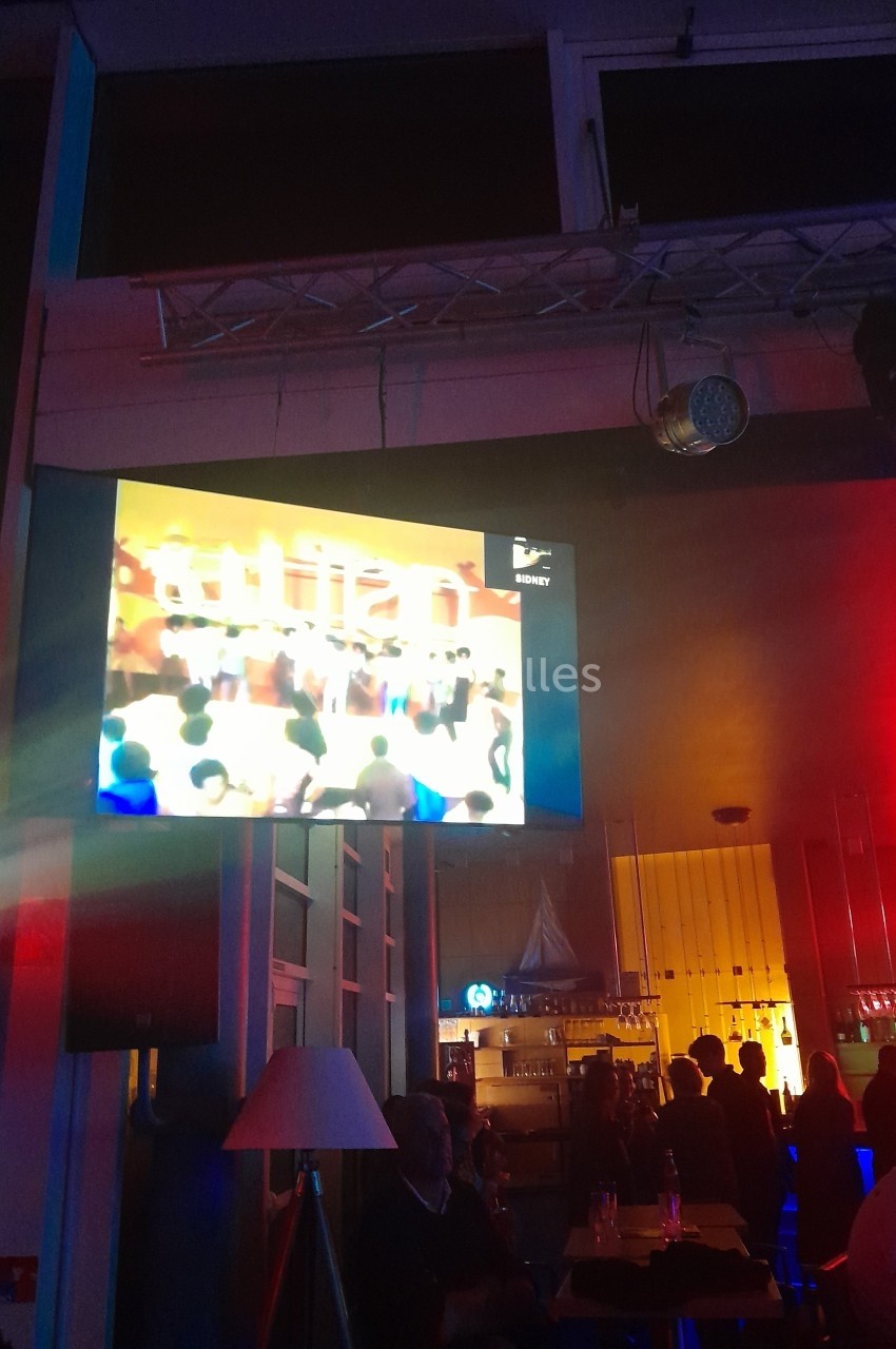 Écran diffusant une émission dans un bar coloré avec des clients assis et un éclairage tamisé.