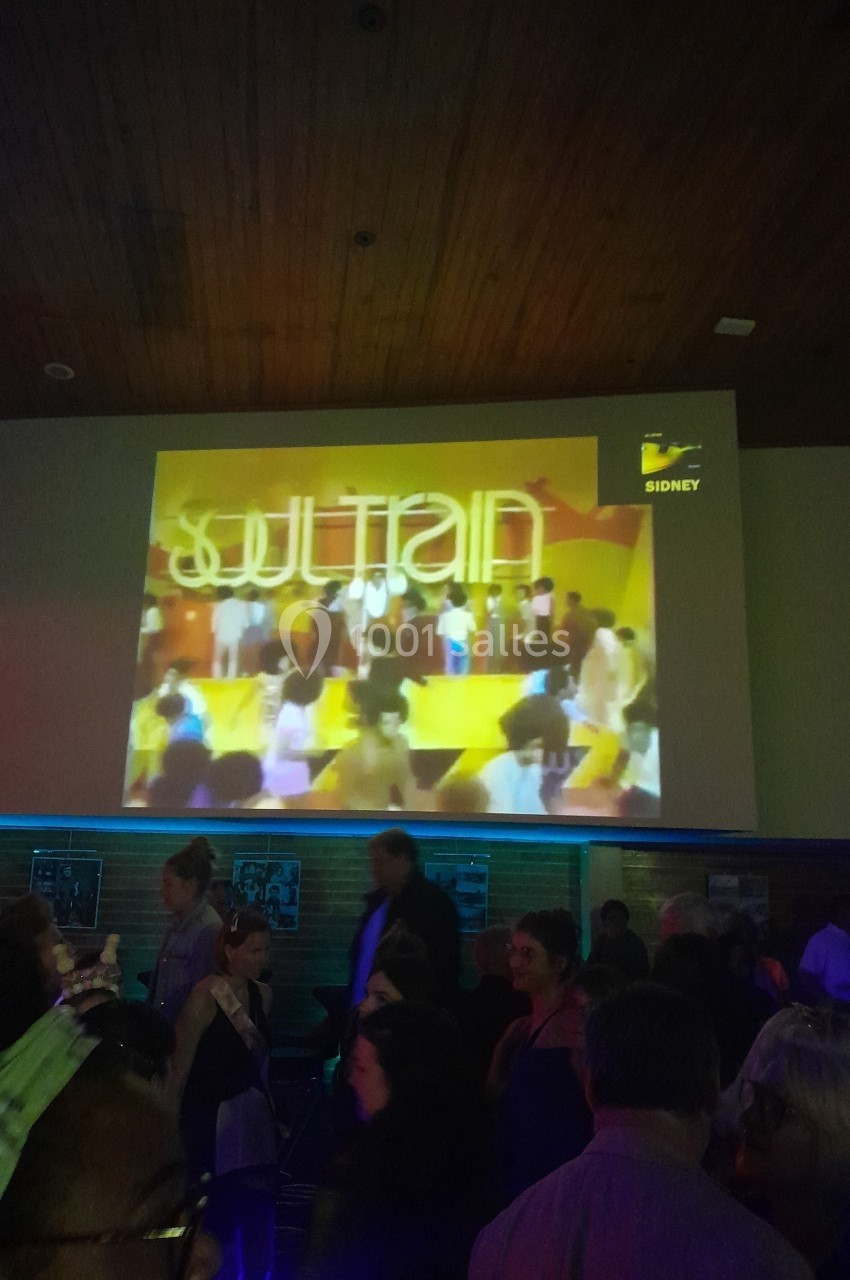 Projection d'une émission rétro sur un écran dans une salle sombre remplie de personnes debout.