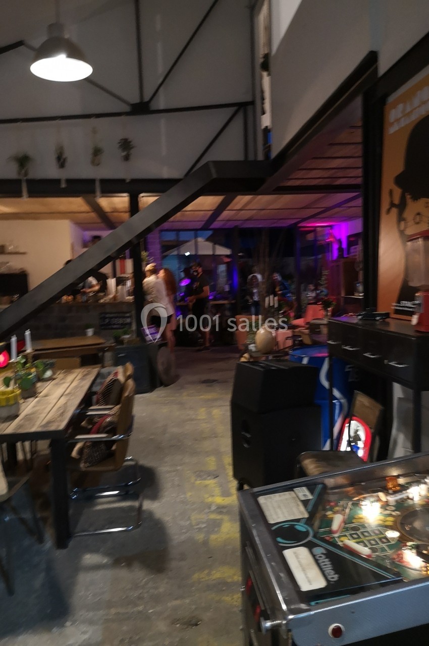 Espace intérieur industriel avec tables, chaises, flipper et décoration lumineuse, ambiance conviviale et décontractée.