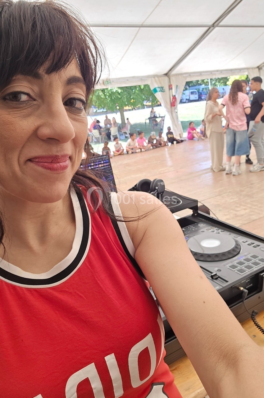Femme souriante en débardeur rouge devant une table de mixage, avec une piste de danse et des danseurs en arrière-plan.