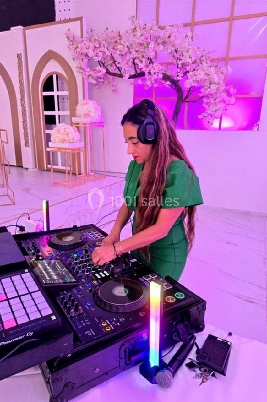 Une DJ en robe verte mixe de la musique sur une table de mixage dans un décor lumineux avec des fleurs de cerisier.