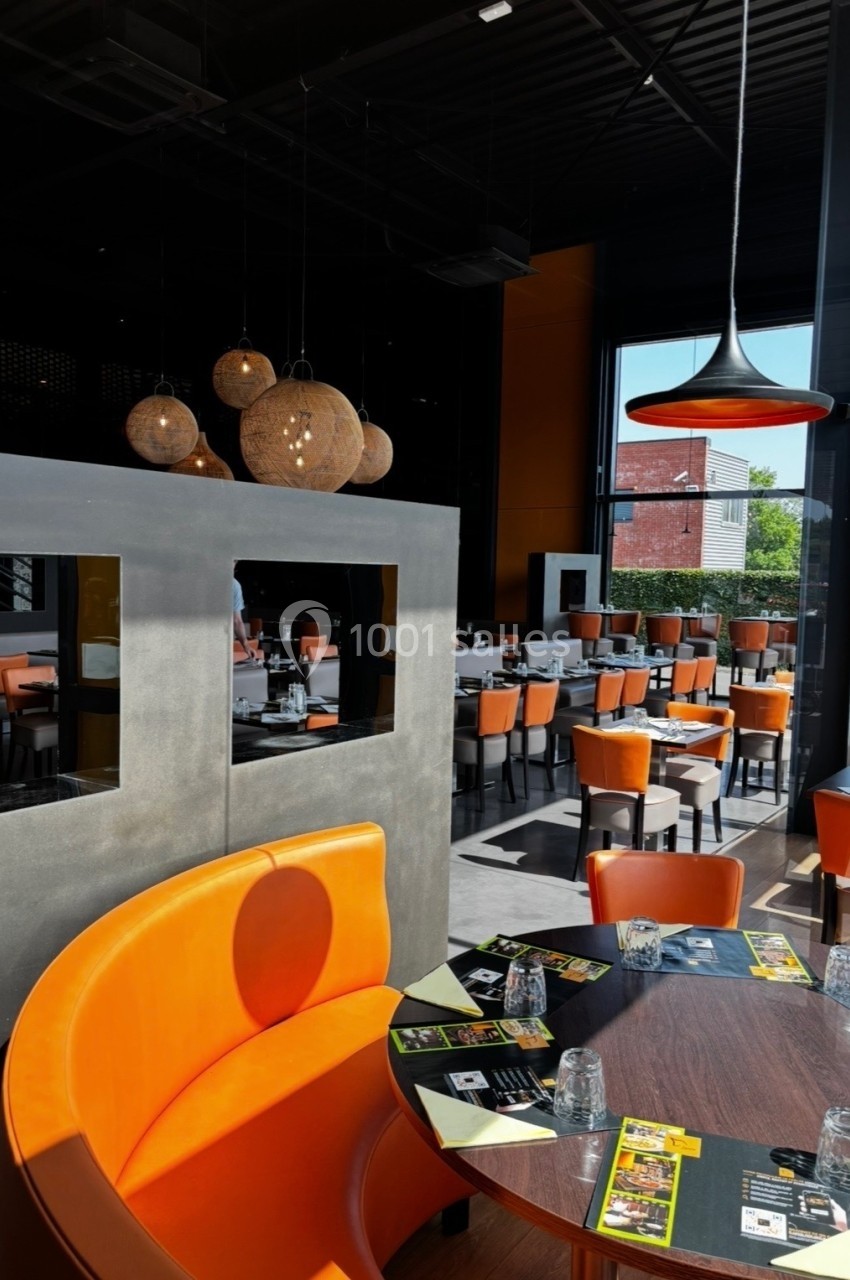 Salle de restaurant moderne avec banquettes orange, tables dressées et grandes baies vitrées laissant entrer la lumière.