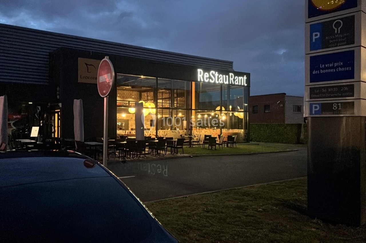 Façade d'un restaurant éclairé en soirée, avec des tables visibles à l'intérieur et un parking à proximité.