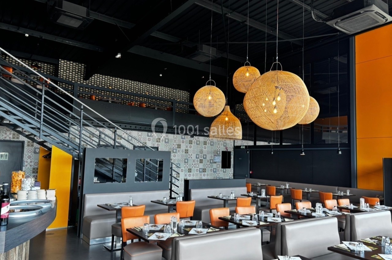 Salle de restaurant moderne avec tables dressées, chaises orange, luminaires suspendus en rotin et escalier métallique.