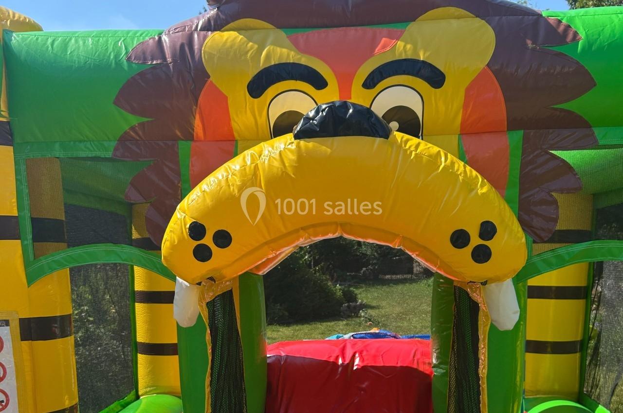 Structure gonflable colorée en forme de tête de lion avec des crocs, située en extérieur sur une pelouse.